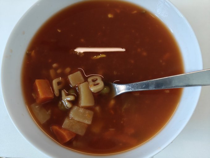 A close-up overhead view of a white bowl filled with dark brown broth soup containing alphabet pasta, diced carrots, potatoes, and green peas, with a metal spoon resting in the bowl.
