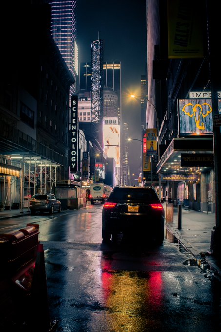 A nighttime street scene in a city, likely New York, with illuminated billboards, a dark SUV driving down a wet road, and the 'Scientology' building visible on the left.