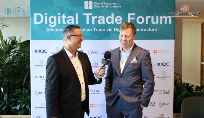 Two men in suits are interviewed in front of a backdrop reading 'Digital Trade Forum', 'Advancing Australian Trade via Digital Infrastructure', and featuring logos for the Digital Economy Council of Australia, XDC, and ATLC, with an 'IMPORTEXPORTG' logo in the corner.