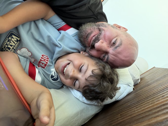 A man and a young boy lie cheek-to-cheek on a bed, both smiling warmly at the camera; the boy wears a gray soccer-style shirt with red stripes.