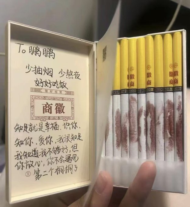 A hand holds open a box of cigarettes with a handwritten note in Chinese addressed to '鵬鵬' expressing care and well-wishes.