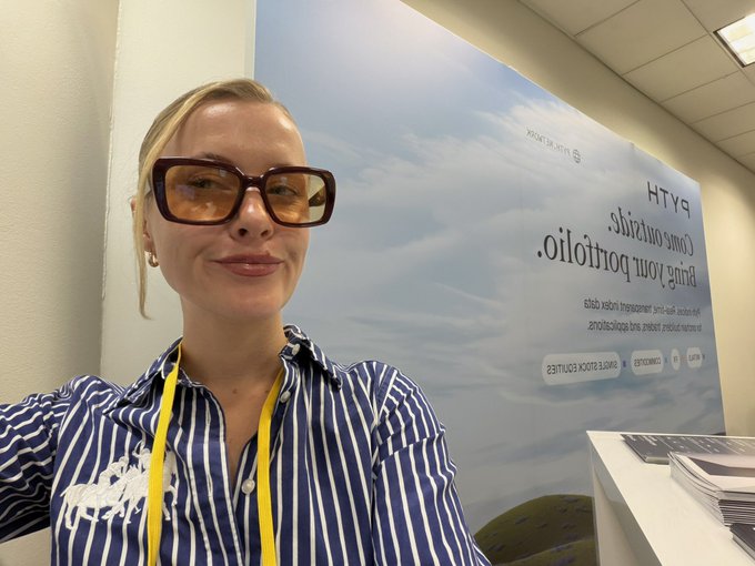 A blonde woman wearing large glasses and a blue and white striped shirt takes a selfie in front of a banner reading 'Bring your portfolio. Come over virtual.', dated 30 September.