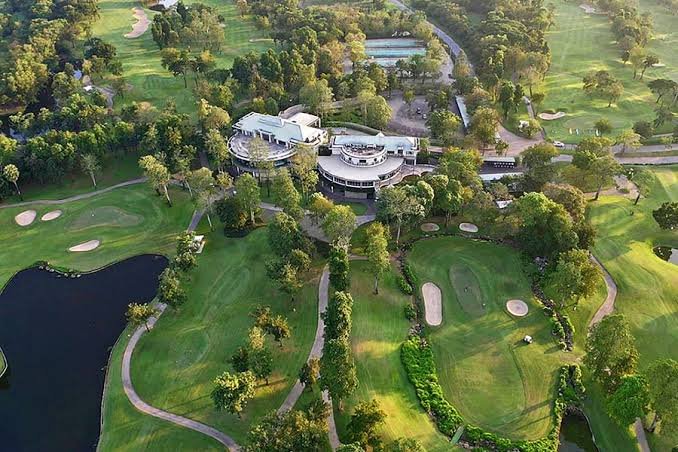 An aerial view of a golf course with a large clubhouse building, surrounded by trees, sand traps, and a small lake.