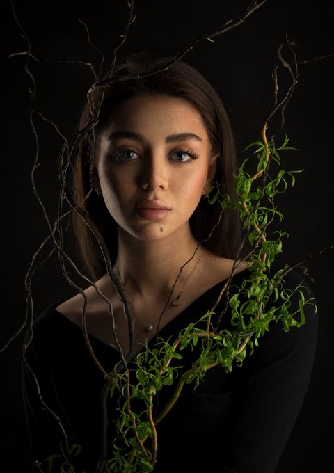 Portrait of a woman with long brown hair surrounded by vines and green leaves against a dark background.