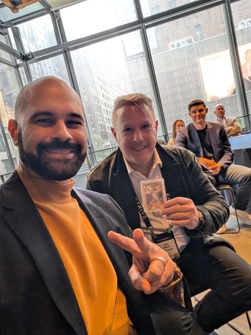 Two smiling men pose for a selfie in a modern glass-walled venue, with the man on the right holding up a Bitcoin-themed card while flashing a peace sign, capturing the camaraderie of a Bitcoin and digital credit gathering mentioned in the post. Other attendees can be seen seated in the background, reflecting the "room full of people taking action" highlighted in the caption.