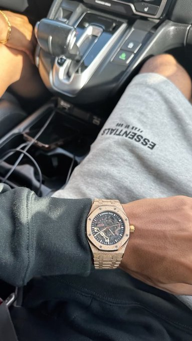 A man in a car shows off his gold Audemars Piguet Royal Oak watch on his wrist, paired with an Essentials grey t-shirt, capturing a "life after larping" flex moment with his driver visible.