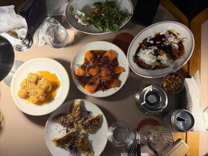 An overhead view of a dining table spread with various dishes including salmon, dumplings, salad, and snacks, along with drinking glasses.