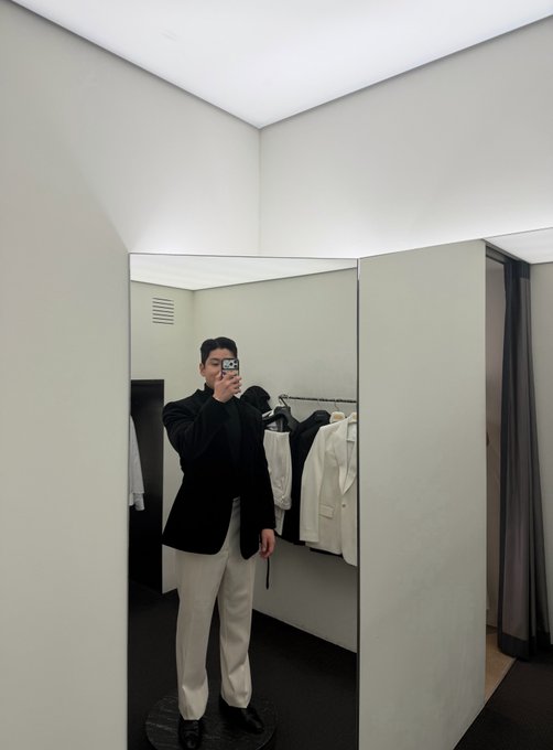 A man in a black blazer and white pants takes a mirror selfie with his phone inside a fitting room with clothing racks visible behind him.