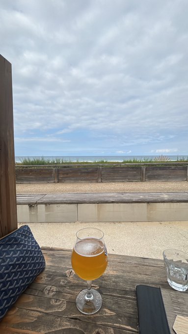 A glass of golden beverage sits on a wooden table overlooking a sandy beach and ocean under a cloudy sky.