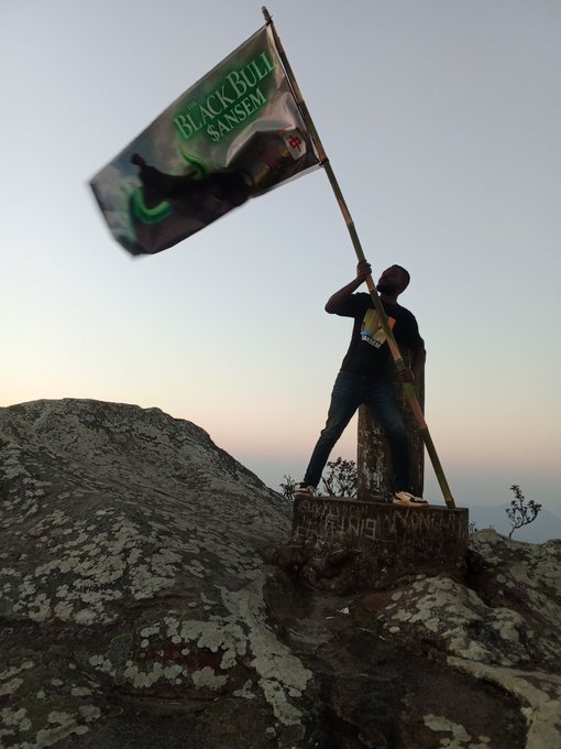 A person standing on a rocky mountain summit at sunset, raising a flag pole with a banner that reads 'BLACKBULL SANSEM'.
