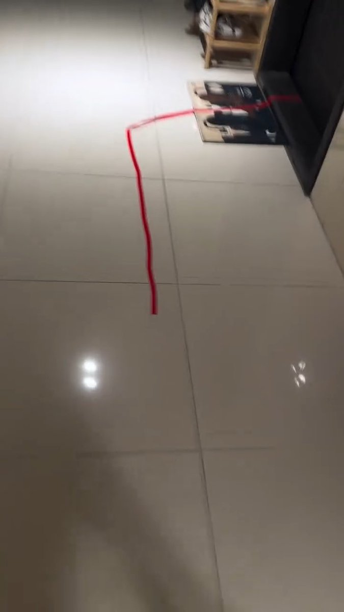 A blurry, low-quality photo of a tiled floor with a red laser line projected across it, extending toward a dark piece of furniture.