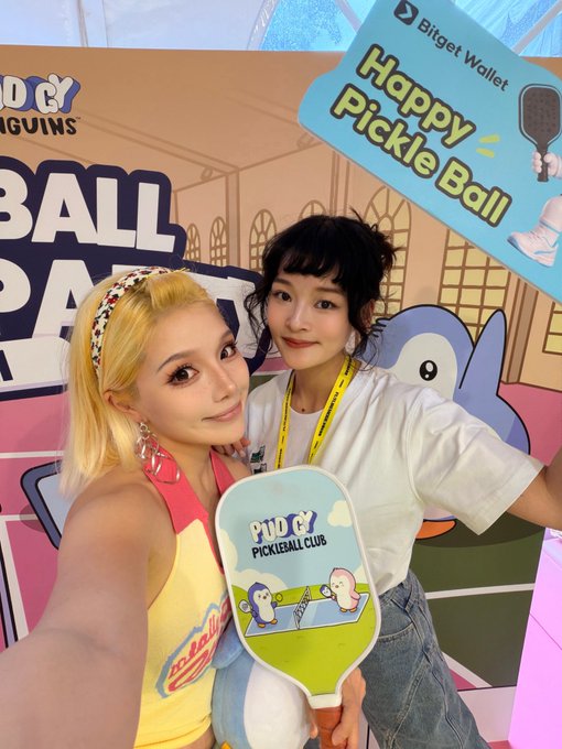 Two friends pose at a Pudgy Penguins Pickleball Social Party, with one holding a "Happy Pickle Ball" paddle branded with the Bitget Wallet logo alongside the Pudgy Penguins mascot. The accompanying post highlights the event and praises how Bitget Wallet's Tiffany blue and white pickleball accessories perfectly match the Pudgy Penguins aesthetic.
