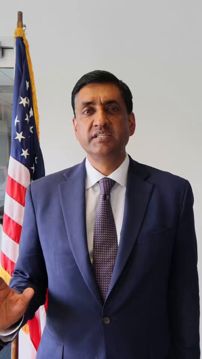 The image features a man dressed in a suit standing next to an American flag, with his hand on his hip. The man appears to be in a professional or formal setting, possibly at work or during a meeting. In the background of the image, there is a post that reads "Debate me instead of threatening lawfare! He should’ve thought about that before demanding Elon be subpoenaed and". This message seems to be directed towards the man standing next to the flag, suggesting some sort of disagreement or conflict between them.