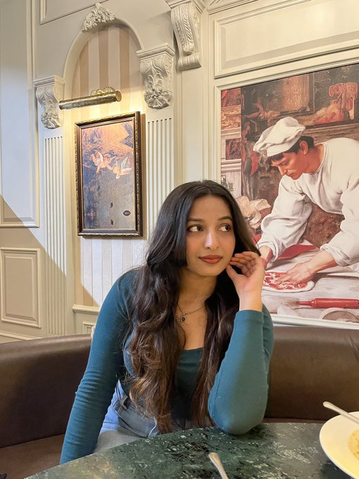 A young woman with long dark wavy hair, wearing a teal long-sleeve top and a necklace, sitting at a table in an ornate restaurant with framed artwork and decorative molding on the walls.