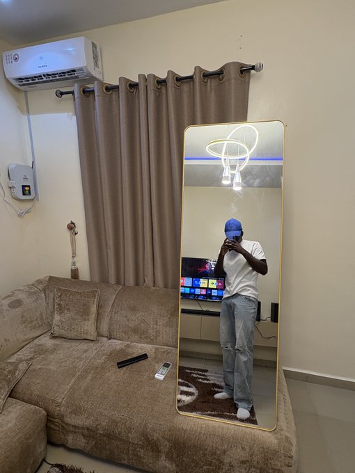A man wearing a blue cap, white t-shirt, and light jeans takes a mirror selfie in his living room, with a TV and decorative lighting visible in the reflection. The casual "TGIF" caption matches the relaxed Friday vibe of the cozy home setting.