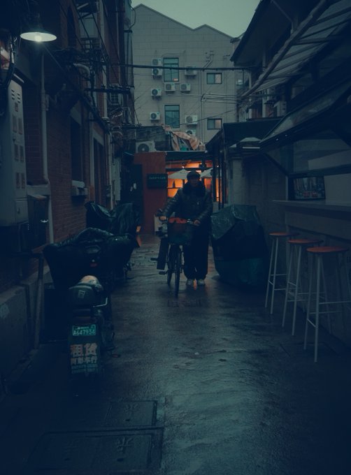 A moody, cinematic photograph of a person walking with a bicycle down a dimly lit, wet alleyway lined with covered motorcycles and a bar stool seating area.