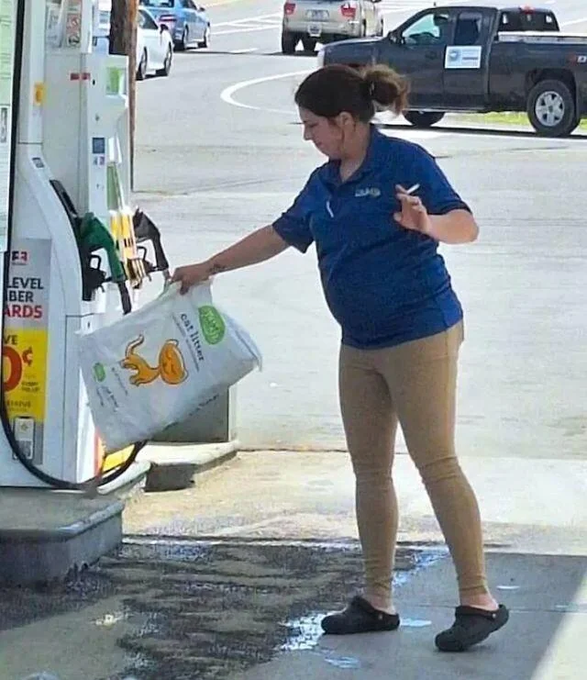 A woman in a blue polo shirt and khaki pants standing at a gas station, holding a plastic bag with a fox logo while smoking a cigarette.