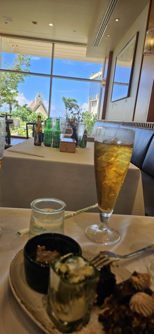 A tall glass of iced tea sits on a restaurant table near large windows, with bottles visible on another table in the background.