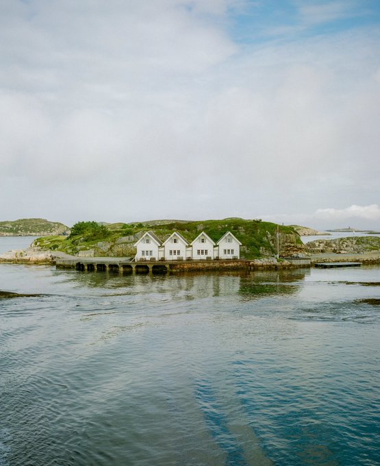 Four charming white cottages stand in a row along the rocky shoreline of a small Norwegian island, their reflections shimmering in the calm blue waters as a ferry passes by under a softly clouded sky.