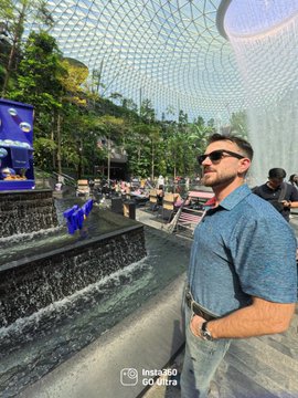 Man wearing sunglasses and a blue polo shirt stands in a modern airport lounge with curved glass architecture and waterfalls in the background.