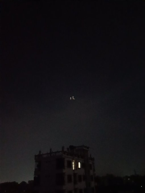 A nighttime photo of a dark sky showing two bright lights (possibly a plane or drone) above a silhouetted building with a lit window.