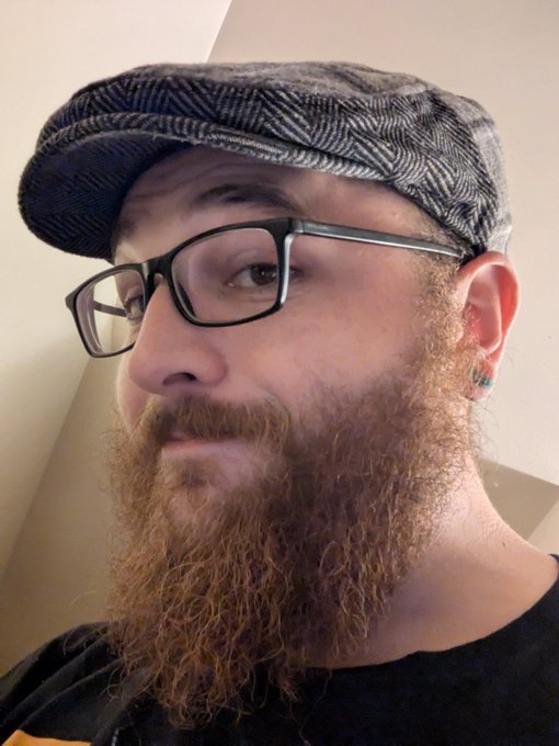 A close-up portrait shows a man with a full reddish-brown beard, wearing black-rimmed glasses and a gray herringbone flat cap, looking thoughtfully to the side.