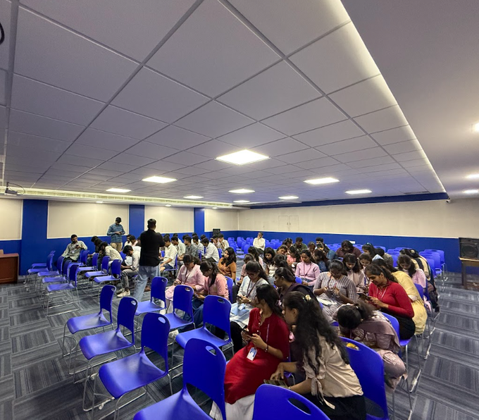 A group of students sits on blue chairs in a large room with white and blue walls, appearing to attend a class or lecture.