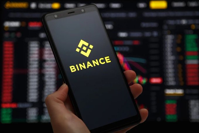 A hand holds a smartphone displaying the yellow Binance logo, with a background of a stock or crypto trading screen showing red and green numbers.