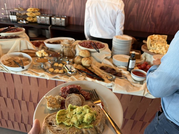 A first-person view shows a white plate holding toast topped with avocado, a small cup of food, and granola with a fork and knife, while a buffet spread featuring pastries, breads, and various dishes fills the background.