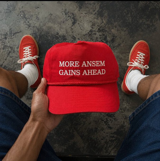 A first-person photo of someone holding a red baseball cap reading 'MORE ANSEM GAINS AHEAD' between their legs, wearing red sneakers and white socks on a gray floor.