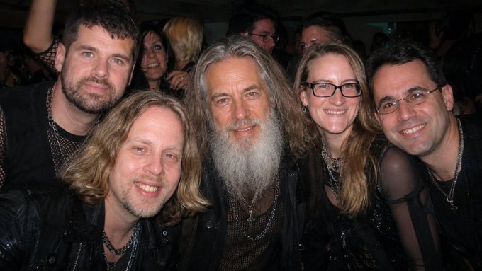 A group of five people, including a man with long gray hair and a beard in the center, smile together for a close-up photo in what appears to be a crowded event setting.