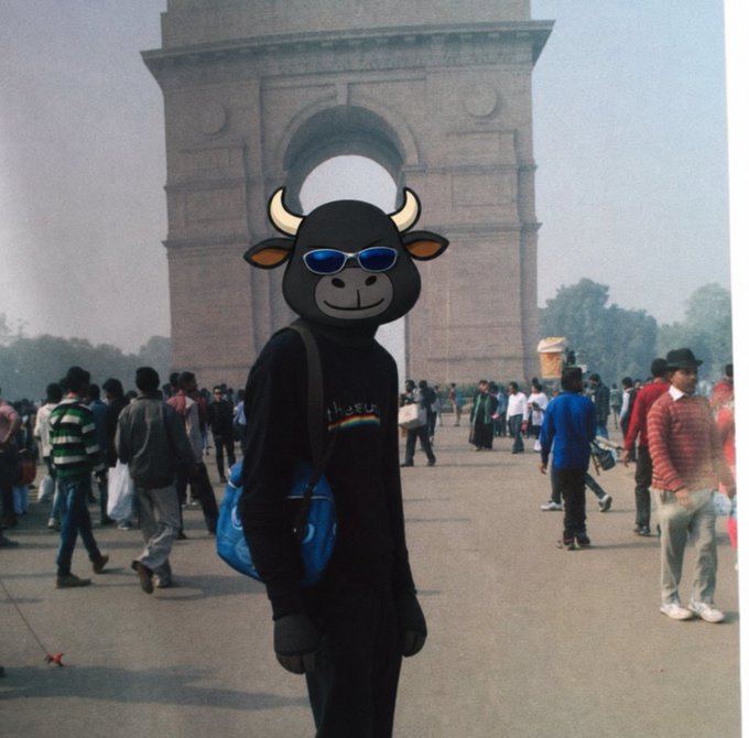 A person in a black outfit wearing a blue shoulder bag stands in front of the India Gate monument in New Delhi, with their face covered by a cartoon bull mask sticker wearing sunglasses.
