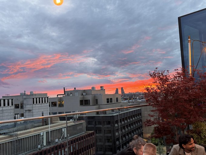 The image shows a rooftop bar with people enjoying drinks, as indicated by the presence of multiple individuals seated at tables. The sky above them has an orange hue, suggesting that the sun is setting or rising. The scene takes place in a cityscape, which adds to the urban atmosphere and highlights the location's appeal for socializing and dining experiences.
