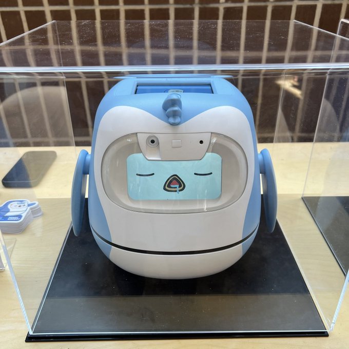 A blue and white robot with a digital face displayed on its screen, enclosed in a clear acrylic display case on a table.