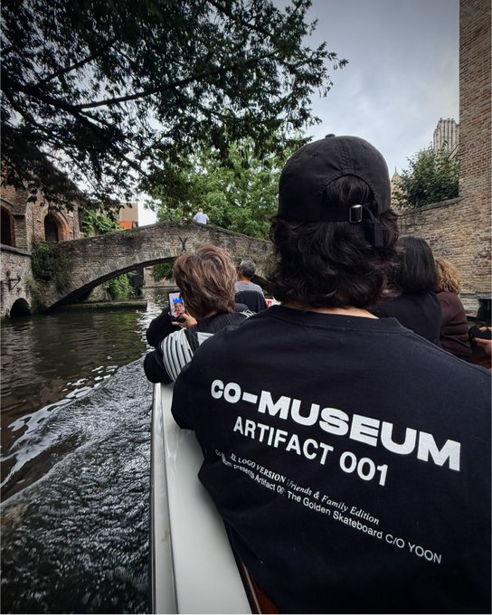 A person wearing a "CO-MUSEUM ARTIFACT 001" t-shirt takes in the view from a boat passing under a stone bridge, capturing the moment on their phone. The scene embodies the post's message of enjoying life's simple pleasures and staying grounded in the journey, even while pushing forward with purpose.