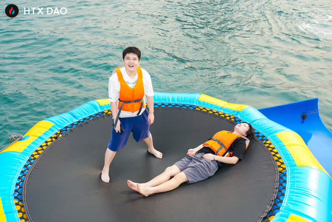 Two people wearing life jackets on a floating trampoline platform on the water, with one person standing and smiling at the camera and the other lying down; the 'HTX DAO' logo is in the top left corner.