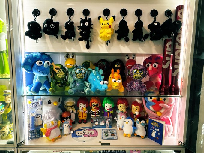 A display case filled with various plush toys, including character mascots, small penguin figures, and orange baseball bat keychains.