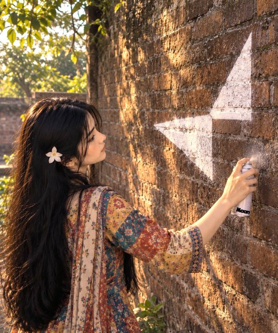 A young woman with long dark hair and a flower accessory stands in profile, spray-painting a white star on a sunlit brick wall while wearing a patterned bohemian dress.