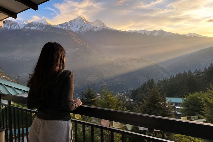 A woman with long dark hair stands on a balcony, gazing out at a breathtaking mountain valley landscape illuminated by golden sunset light.