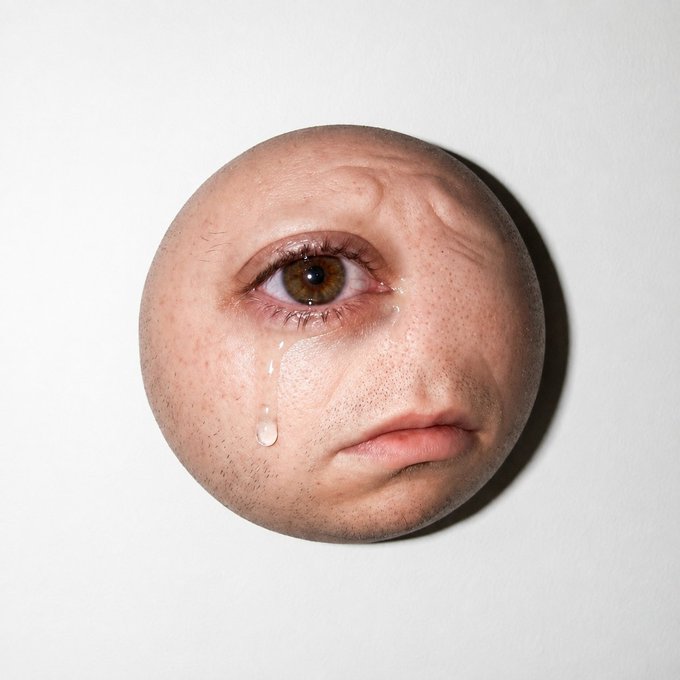 A surreal circular artwork showing a partial human face with a single tear rolling down the cheek.