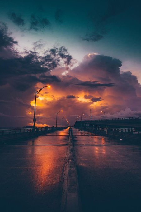 Dramatic sunset photograph over a wet, empty bridge lined with glowing streetlamps under a sky of deep orange and purple clouds.