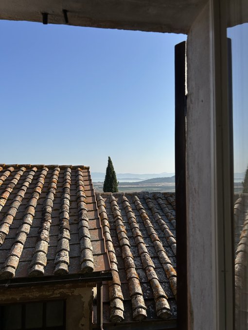 A view through a window frame showing terracotta tile rooftops, a tall cypress tree, and rolling hills under a clear blue sky.