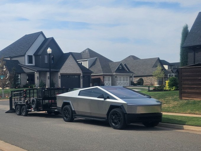 A Tesla Cybertruck in stainless steel parked on a suburban street in front of large houses, towing a black trailer loaded with off-road vehicles.