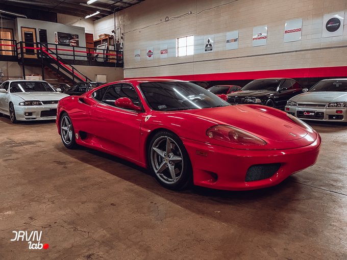 A red Ferrari 360 Modena is parked in a garage showroom with other vehicles, including what appears to be a Nissan Skyline, with various automotive brand signs displayed on the wall.