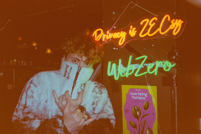 A person wearing a fuzzy blue and white jacket poses in front of neon signs reading 'Privacy is ZEC99' and 'WebZero,' next to a purple poster featuring a head illustration.