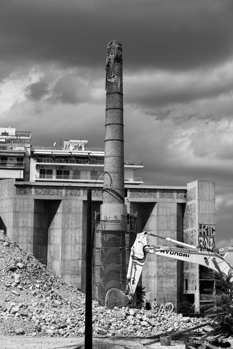 A black and white photograph of a tall industrial chimney with graffiti, set against a dramatic cloudy sky, with a Hyundai excavator and rubble visible in the foreground.