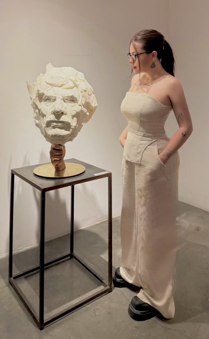 A woman in a beige strapless top and wide-leg pants stands in a gallery next to a sculpted white head with rough, textured features displayed on a black pedestal.