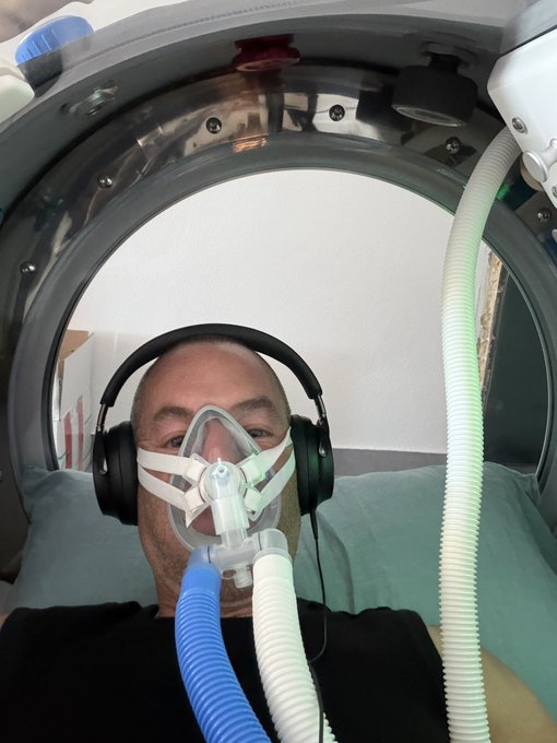 A man wearing headphones and a breathing mask lies inside a hyperbaric oxygen chamber, with hoses delivering air to him. The image illustrates the post's message about prioritizing health investments—like a hyperbaric chamber purchased with crypto—over luxury items such as cars, watches, and yachts.