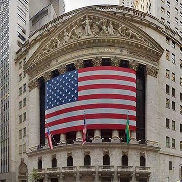 The image features a large American flag hanging from the top of a building, possibly the New York Stock Exchange (NYSE). The text "BREAKING: RIPPLE’S U.S. NATIONAL BANK LICENSE CONFIRMED AS “IMMINENT.”" is present in the image, which suggests that the bank's license has been recently confirmed by the SEC. This news can be considered a significant event for the NYSE and its stakeholders as it indicates regulatory approval of the bank's operations and financial stability.