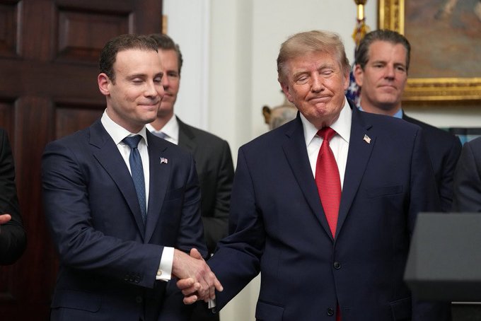 Two men in dark suits shake hands and smile in an ornate room, with one wearing a red tie and an American flag pin while the other wears a dark tie and a small pin. The setting features wood-paneled walls and gold-framed artwork, suggesting a formal government venue. The image's connection to the post about the CFTC sending new crypto rules to the White House implies the handshake may symbolize a regulatory milestone, possibly involving crypto policy collaboration, though no specific text, numbers, or names are visible in the image to directly confirm the identities or exact event depicted.